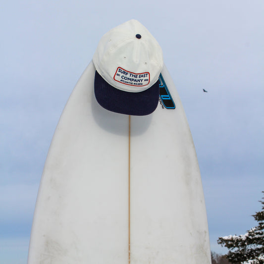 A white and navy blue cap with Surf the East Company, Atlantic Raised embroidered on it sitting on a white surboard.