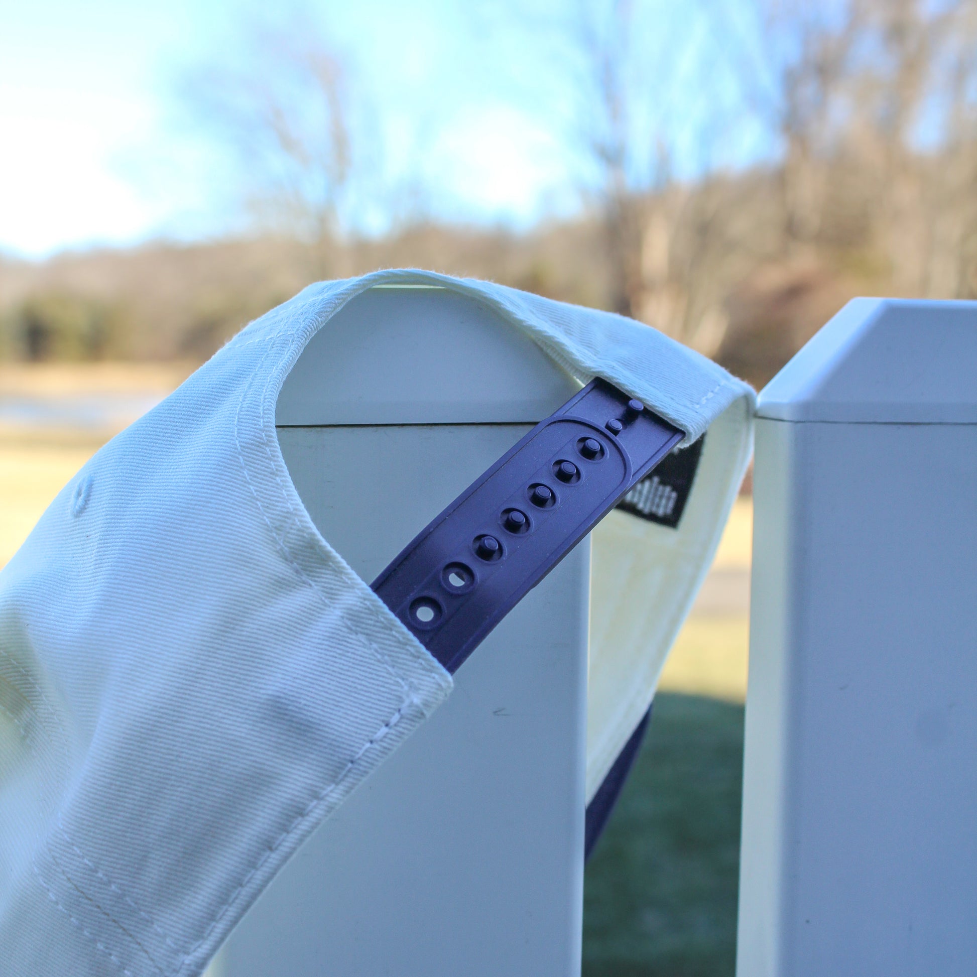 Close-up of the back of a light-colored baseball cap with a navy snapback strap draped over a white fence, with a softly blurred outdoor landscape in the background.