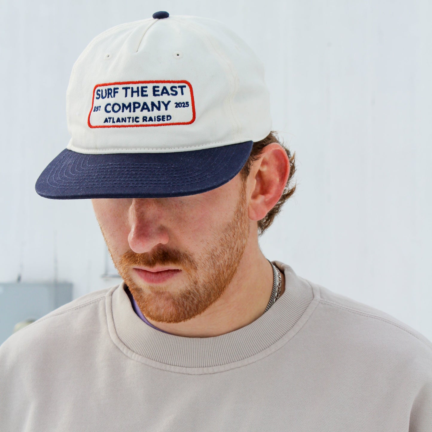 Person wearing a white and navy blue cap with Surf the East Company, Atlantic Raised embroidered on it, and a beige sweatshirt.