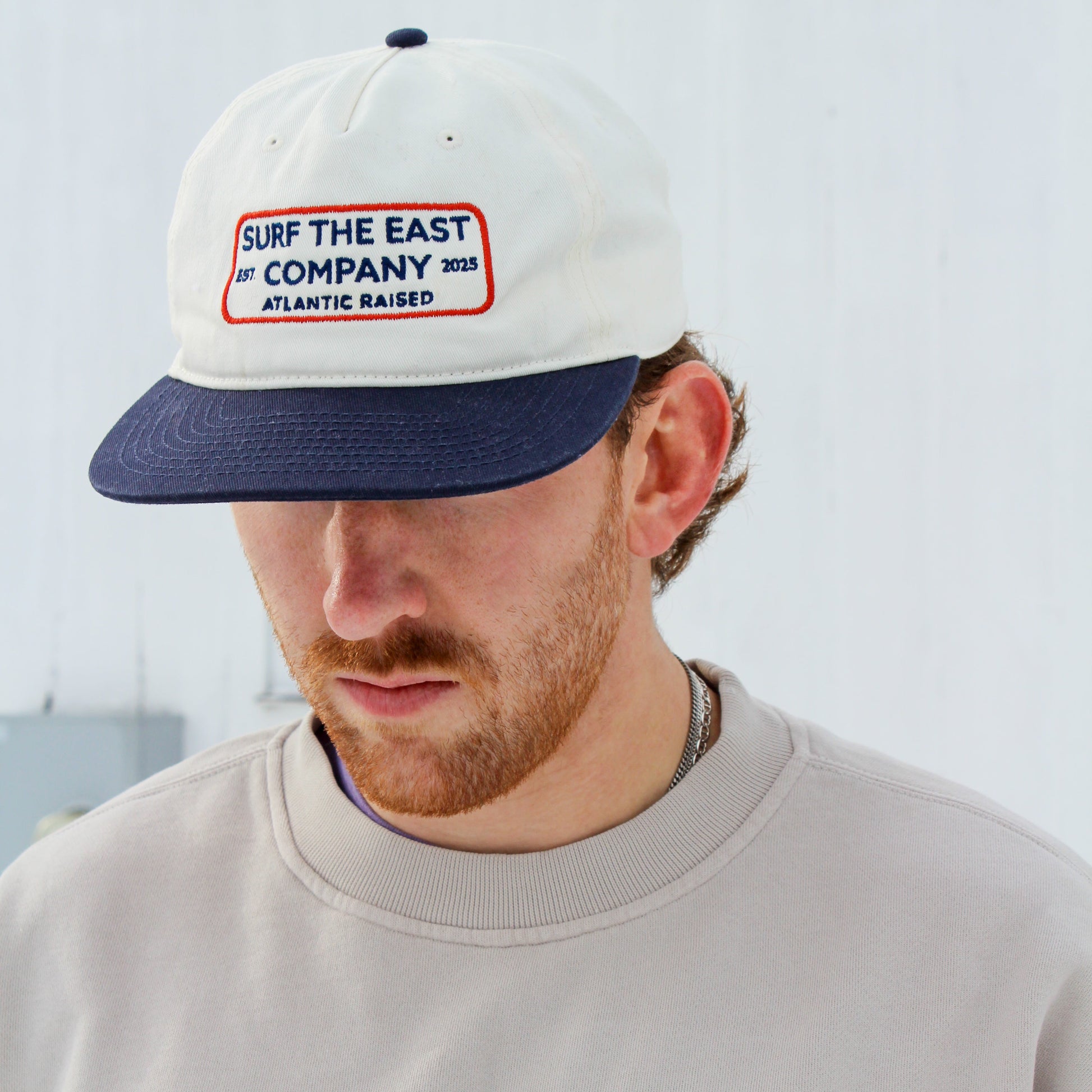Person wearing a white and navy blue cap with Surf the East Company, Atlantic Raised embroidered on it, and a beige sweatshirt.