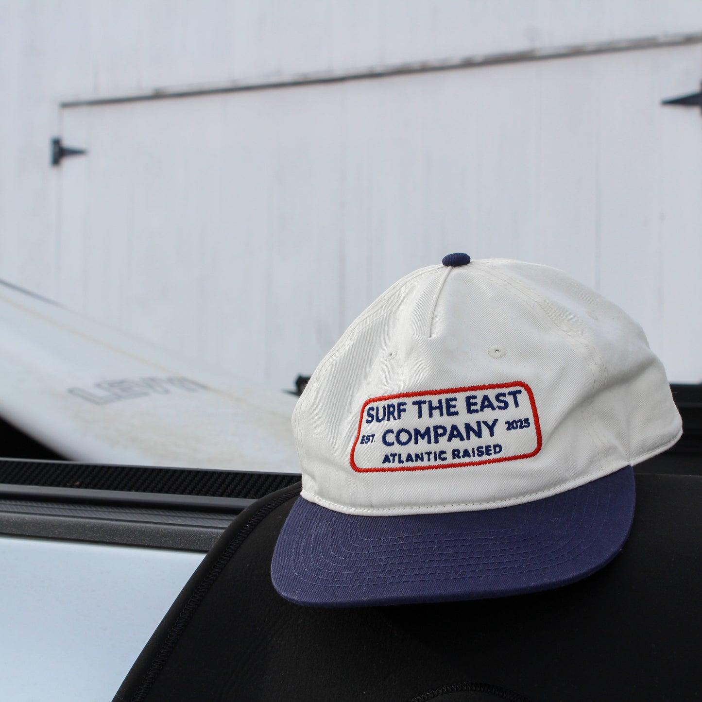 A white and navy blue cap with Surf the East Company, Atlantic Raised embroidered on it, sitting on the edge of a truck with a surfboard in the background.