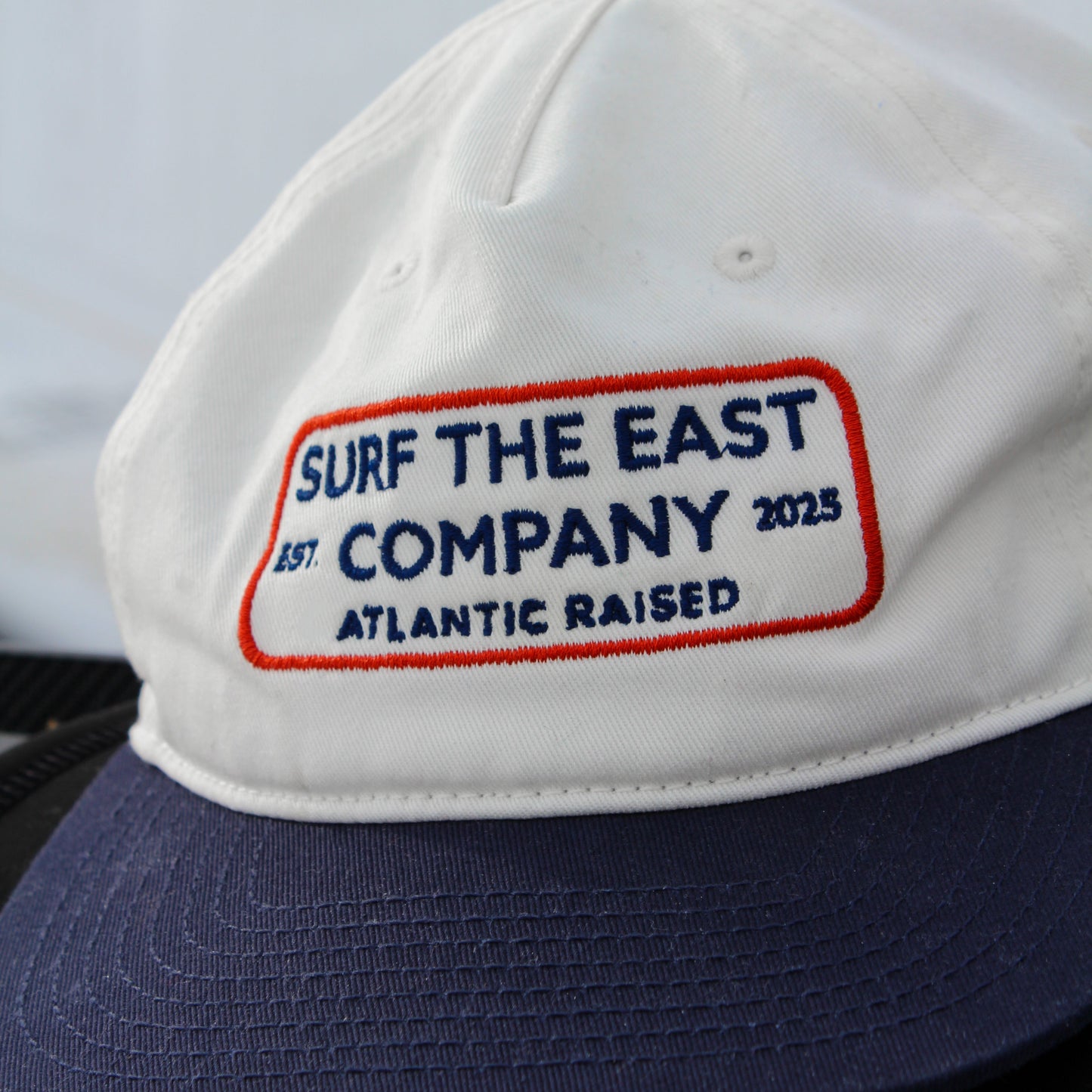A white and navy blue cap with Surf the East Company, Atlantic Raised embroidered on it.