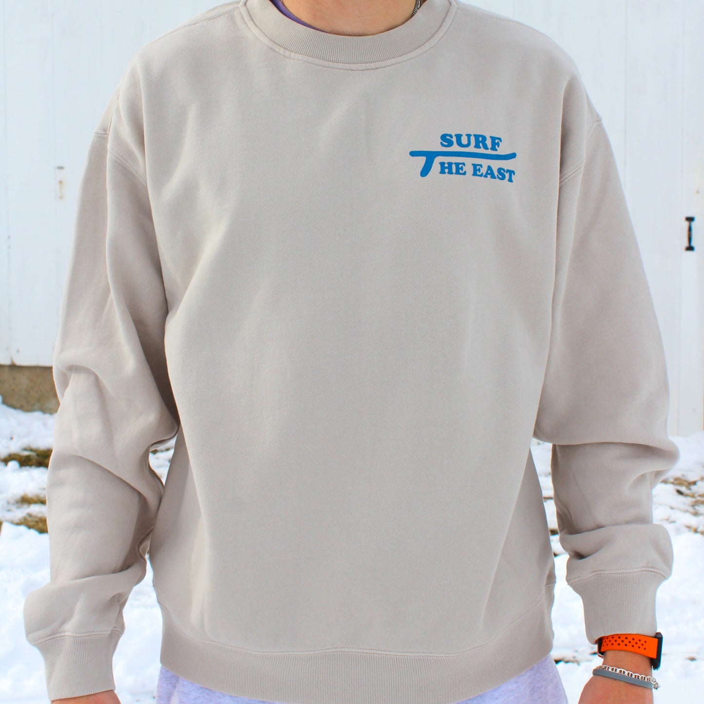 Man wearing beige sweatshirt with blue Surf the East wording on it, standing in front of a white barn with snow on the ground.