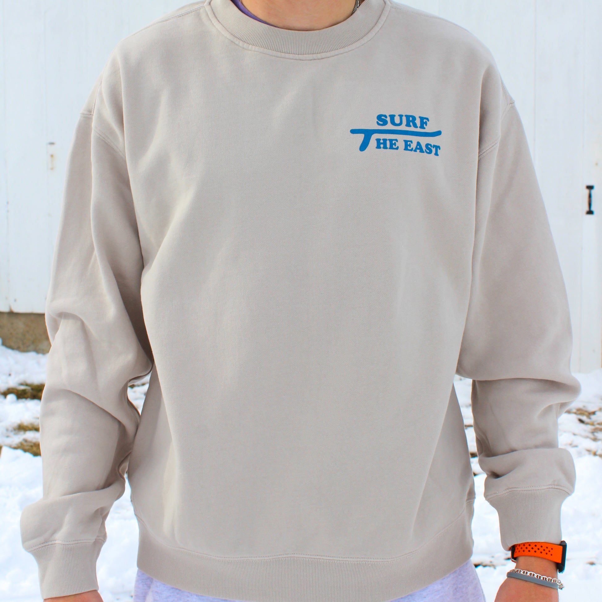Man wearing beige sweatshirt with blue Surf the East wording on it, standing in front of a white barn with snow on the ground.