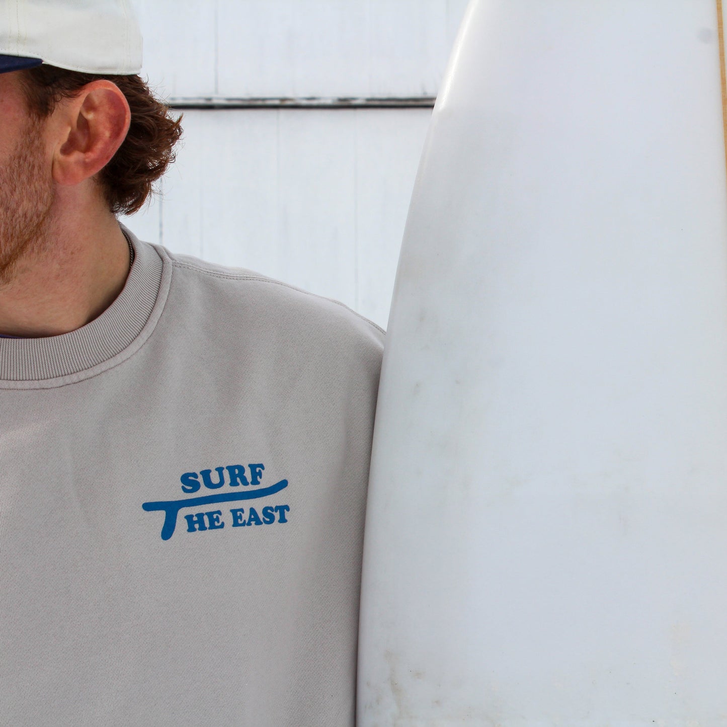 Man wearing beige sweatshirt with blue Surf the East wording on it, standing in front of a white barn with a surfboard next to him.